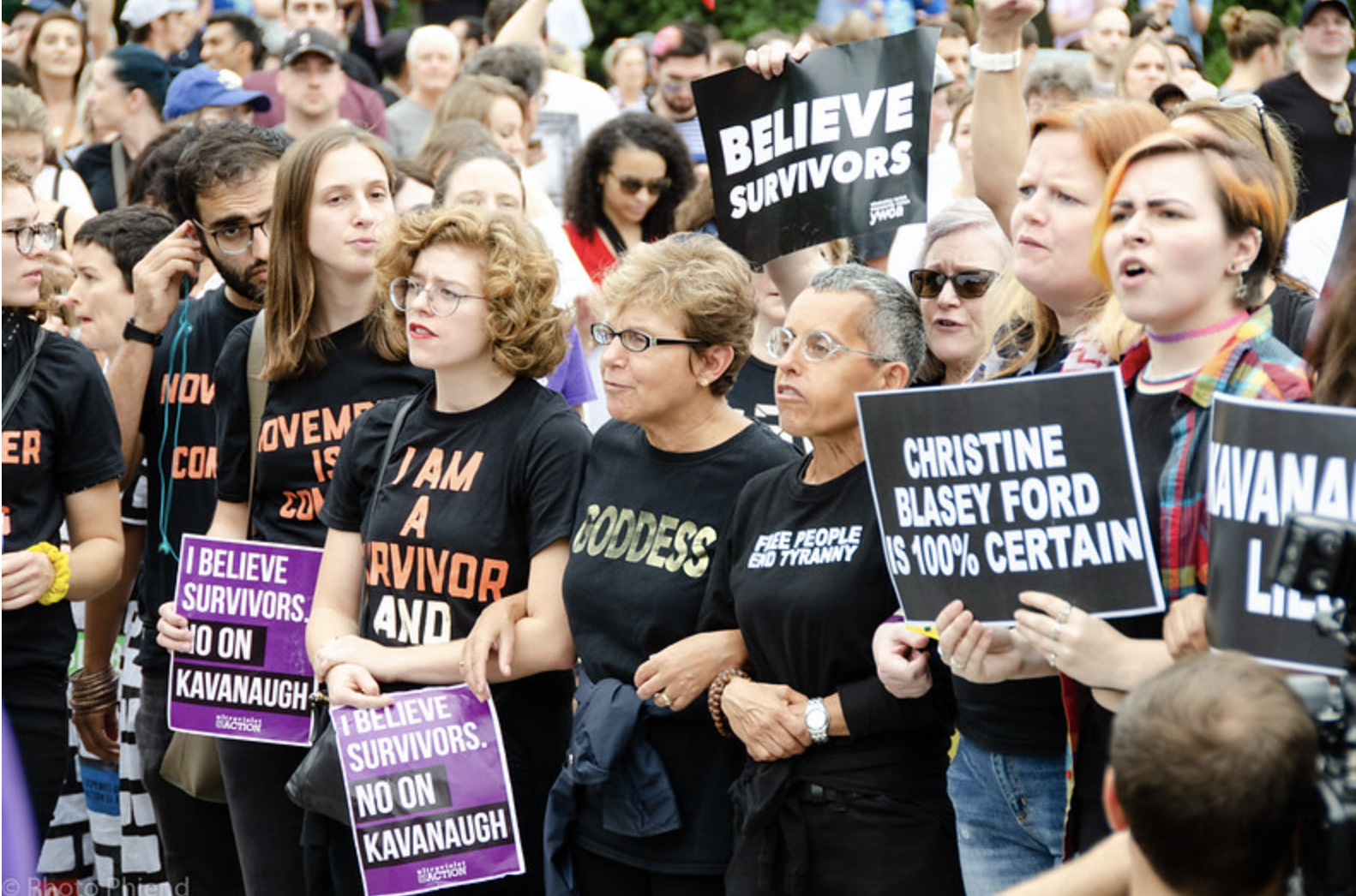 Protesters against nomination of Justice Kavanaugh—many in black T‑shirts—link arms and chant while holding signs like “BELIEVE SURVIVORS,” “I BELIEVE SURVIVORS, NO ON KAVANAUGH,” and “CHRISTINE BLASEY FORD IS 100% CERTAIN.”